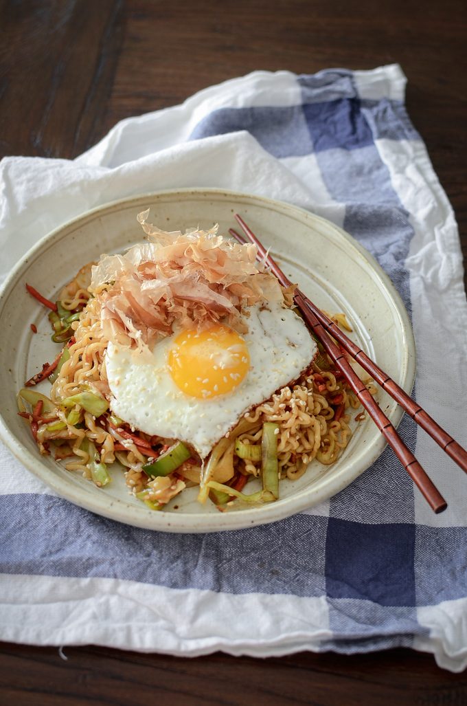 Short ramen and vegetable stir-fry is topped with fried egg and bonito flakes in a bowl.