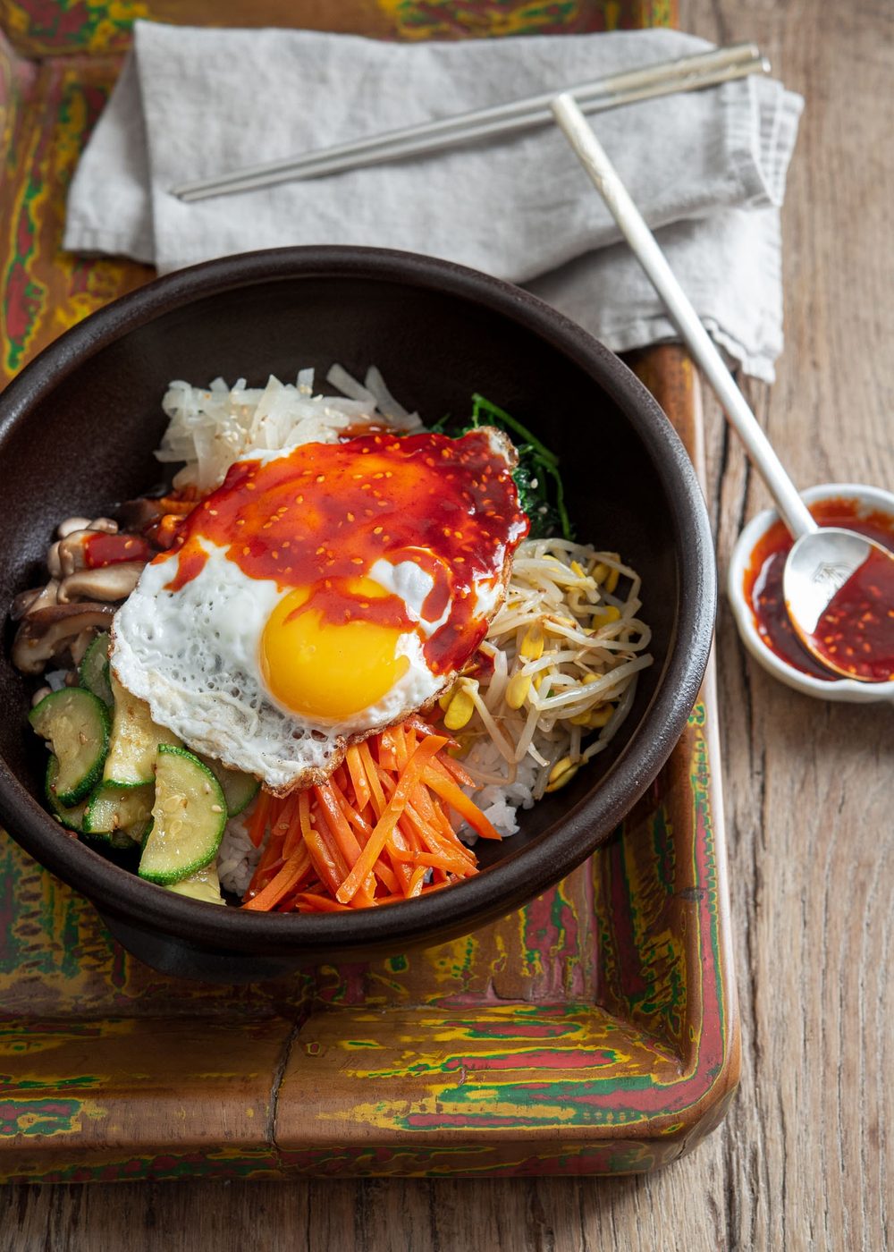 A bowl of Korean bibimbap is on a wooden tray with gochujang sauce on the side