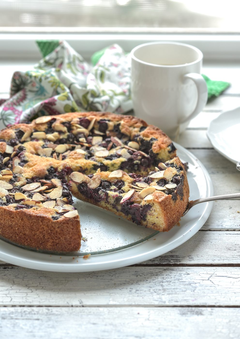 A slice of blueberry cake shows the burst of fresh blueberries on a white plate.