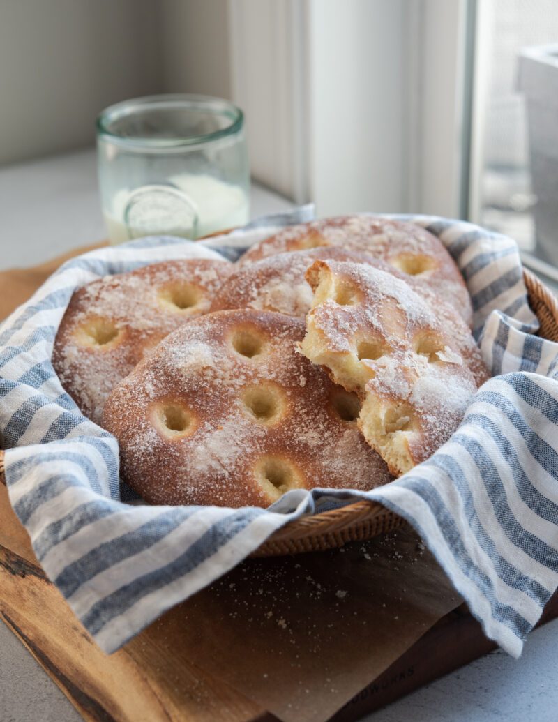 A basket lined with blue stripe napkin is filled with sugary brioche tarts and served with a glass of milk.