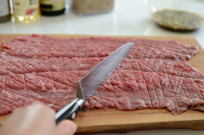 A back of knife is pounding on a thin slice of beef on the cutting board.