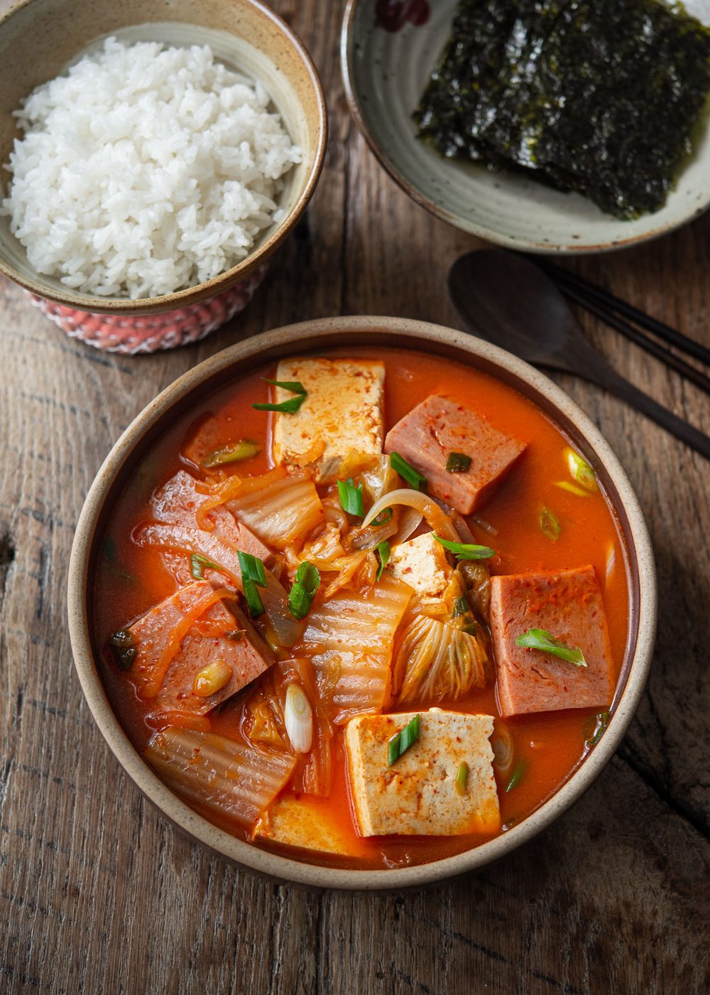 A bowl of kimchi jjigae made with Spam is served with rice and roasted seaweed.