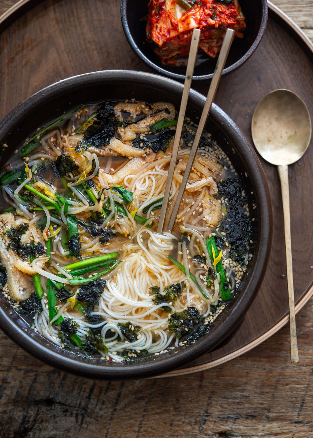 Chopsticks mixing somyeon noodles with tofu, sprouts, and chives in a light Korean broth
