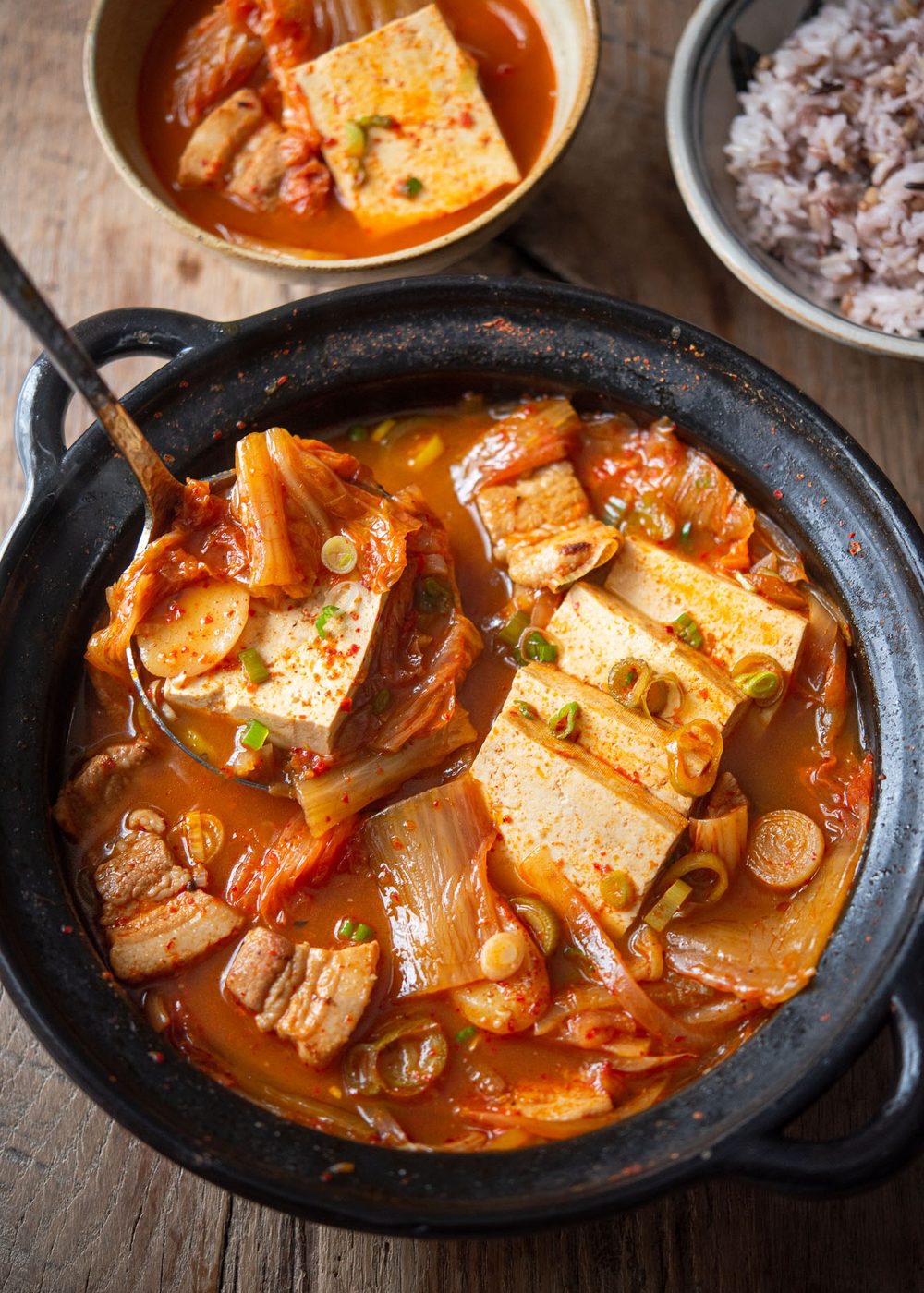 Ladle lifting kimchi jjigae with tofu and pork belly from a simmering pot.