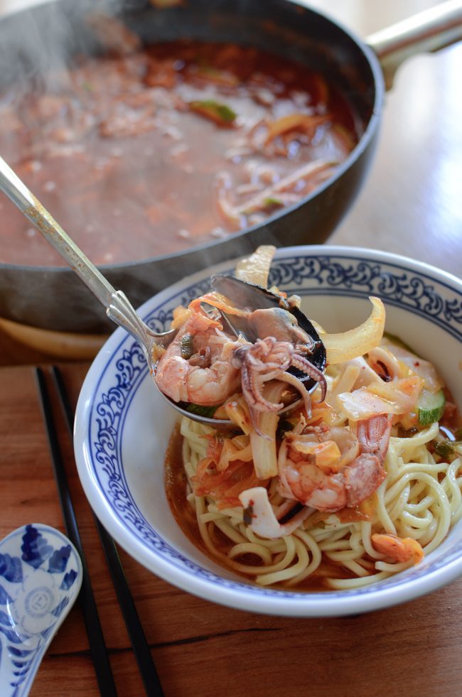 A ladle of spicy seafood soup is poured on the noodles in a bowl.