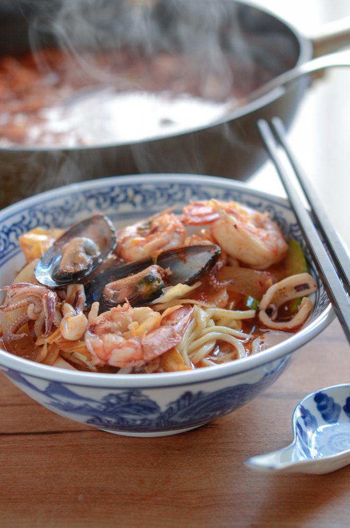 A bowl of noodles are topped with mussel, shrimp, and squid in the spicy soup broth.