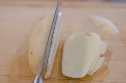 A peeled potato is thinly sliced with a knife.