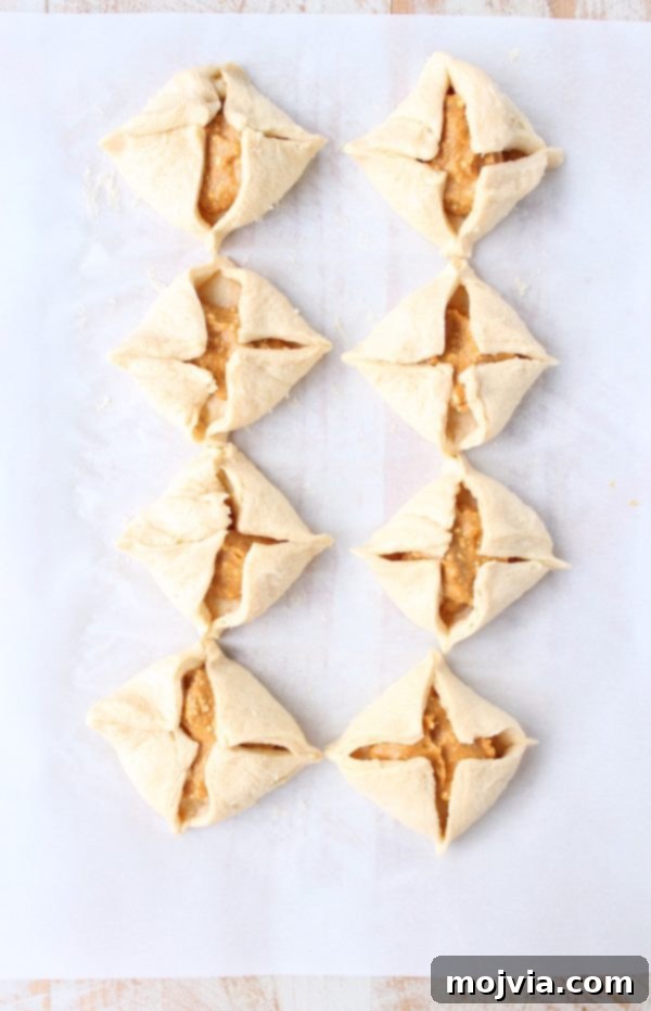 Spiced Pumpkin Pockets 5 Uncooked pumpkin pastries, neatly folded with their corners meeting in the middle, arranged on parchment paper.