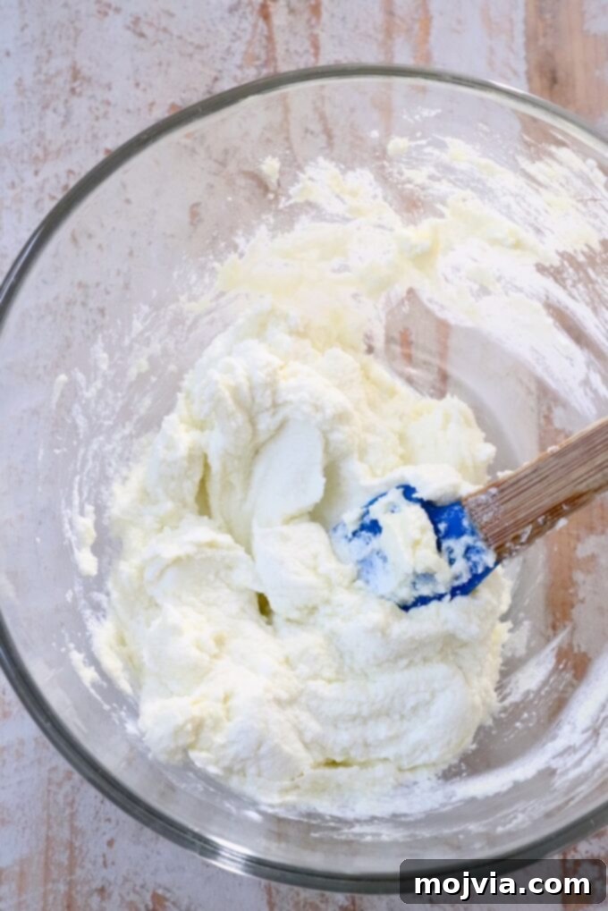 Strawberry Bliss Bites 6 Creamy goat cheese being whipped to a smooth consistency in a clear glass bowl using a blue silicone spatula, ready to be spread on the toasted baguette.