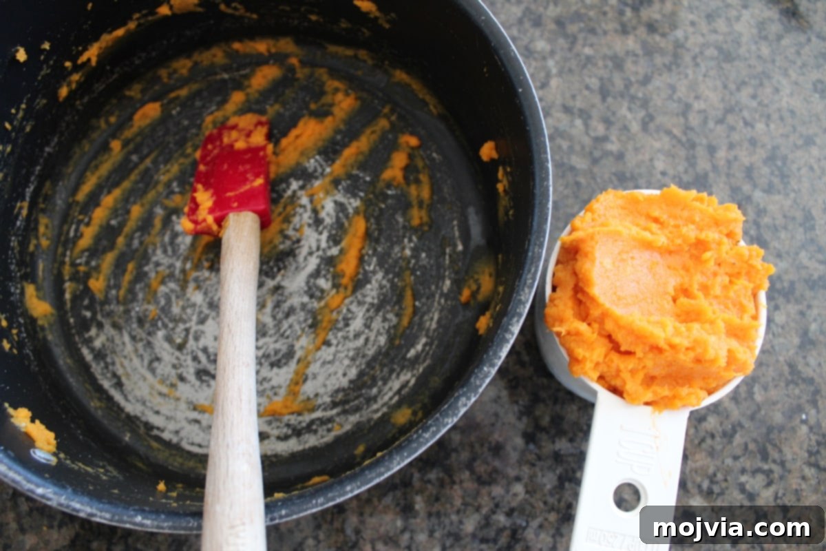 A white measuring cup filled with smooth mashed sweet potatoes, with an empty pot in the background, illustrating the preparation stage.