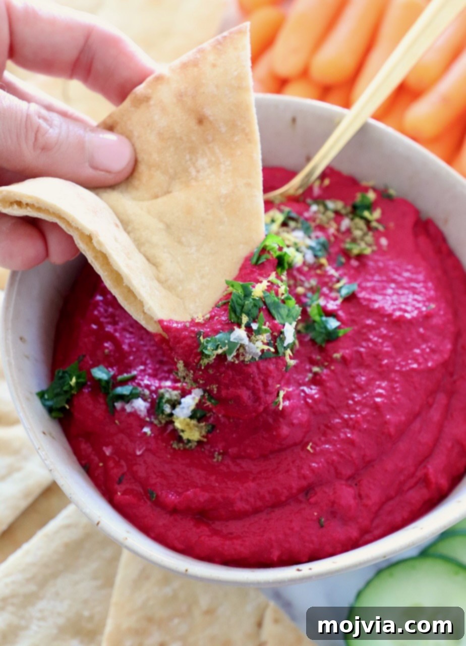Blush Beet Hummus 2 Beet hummus in a bowl with a piece of pita bread dipped into it, showcasing its creamy texture and vibrant pink color.