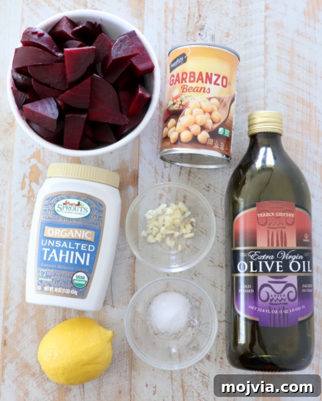 Blush Beet Hummus 3 All ingredients for beet hummus arranged on a white wooden board, including fresh beets, canned chickpeas, tahini, garlic, lemon, salt, and olive oil.