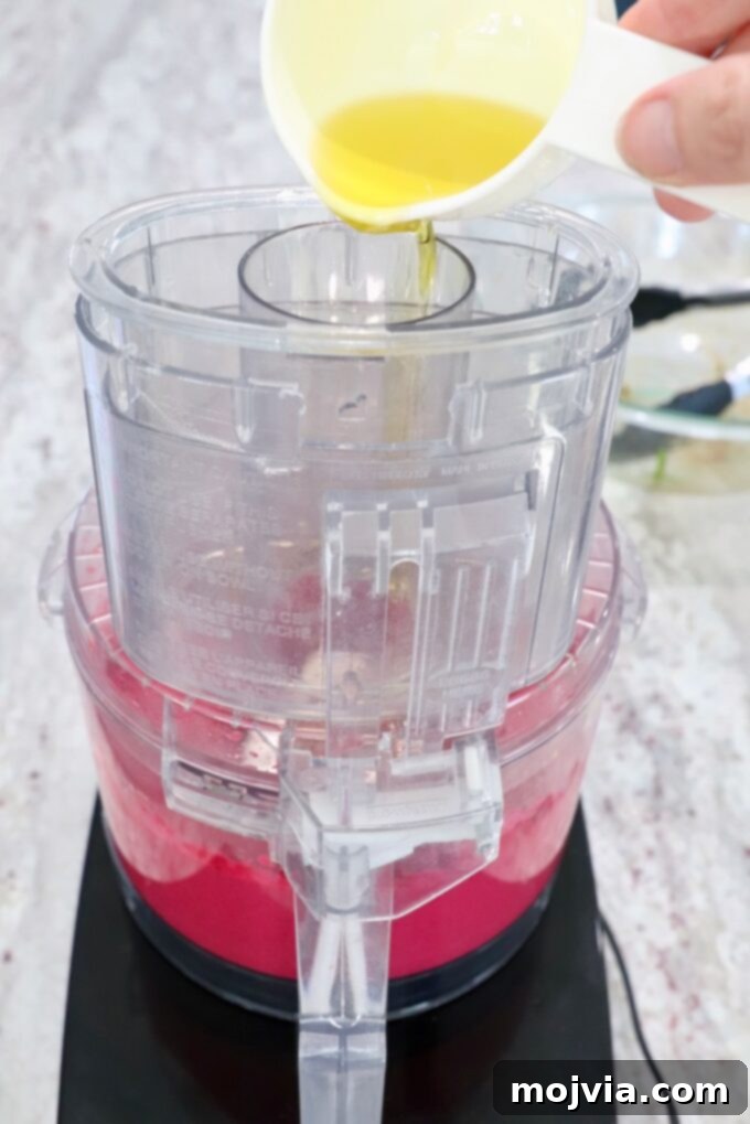 Blush Beet Hummus 9 Olive oil being slowly drizzled into the opening of a food processor lid while beet hummus is blending, to achieve a smooth texture.
