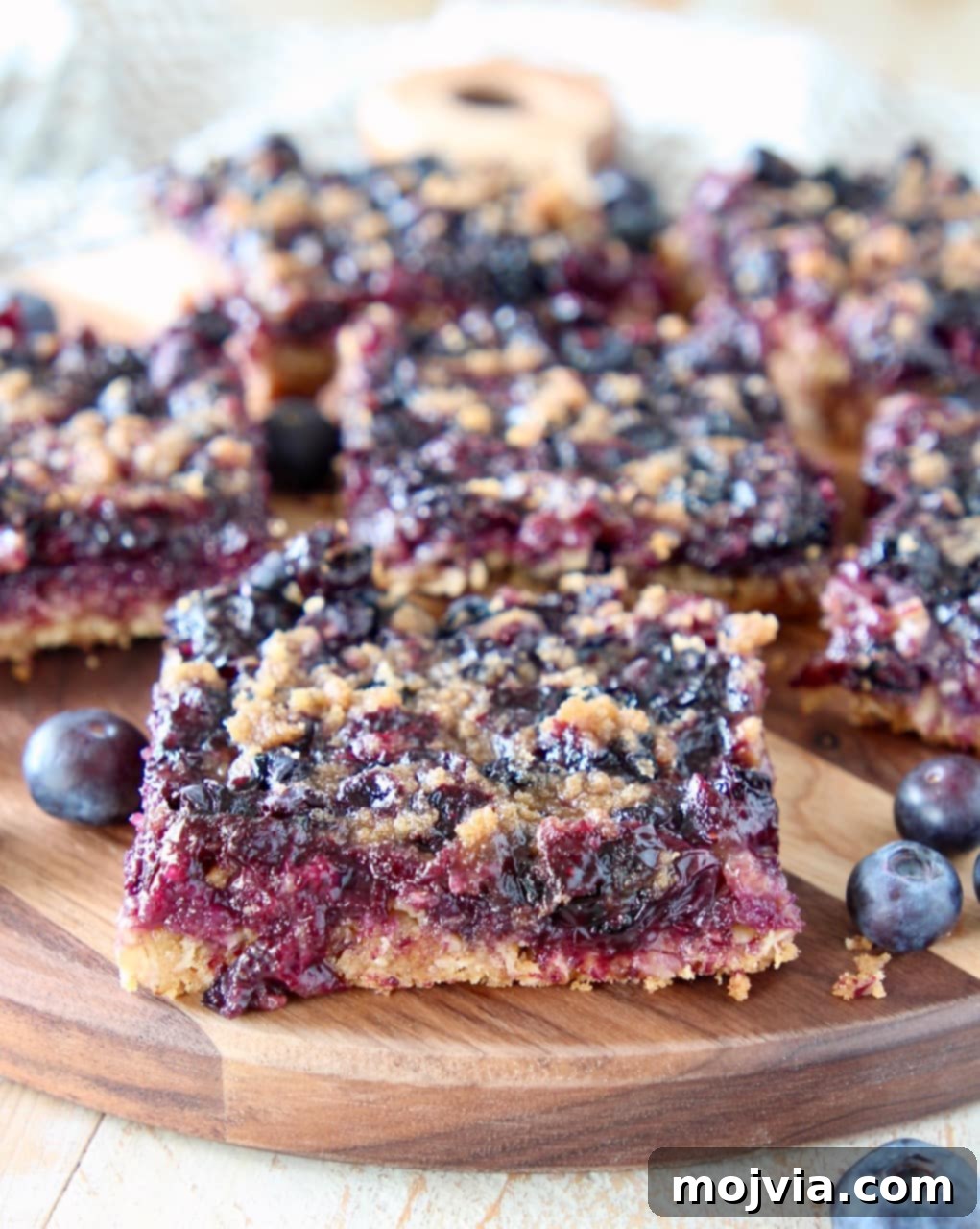 Gluten-Free Blueberry Bliss Bars 2 Beautifully cut gluten-free blueberry oatmeal bars on a wooden cutting board, surrounded by fresh blueberries.