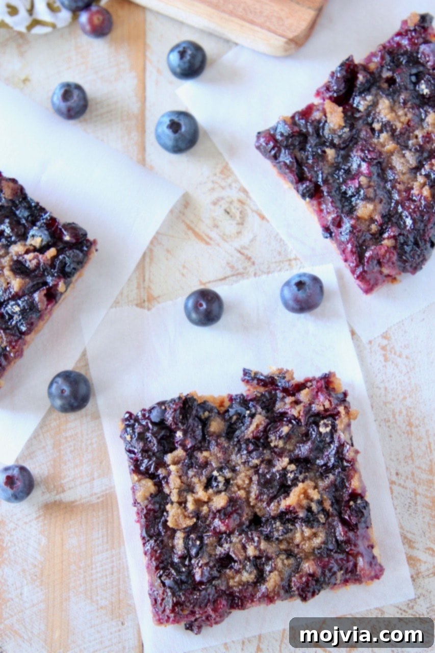 Gluten-Free Blueberry Bliss Bars 3 Overhead shot of freshly baked gluten-free blueberry oat bars, cut into neat squares, resting on white parchment paper with scattered fresh blueberries.