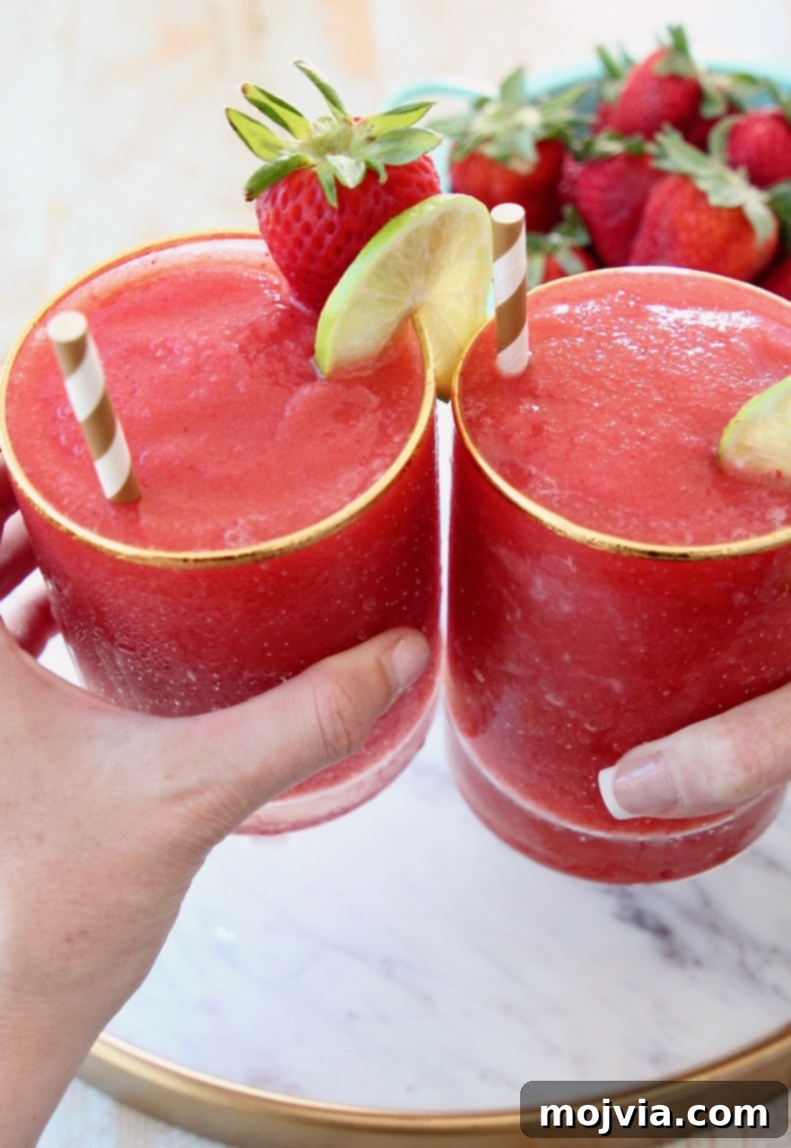 Basil Infused Strawberry Daiquiri 3 Two hands holding elegant glasses of frozen strawberry daiquiris, adorned with gold and white striped straws and fresh lime wedges, set against a bright, inviting backdrop.
