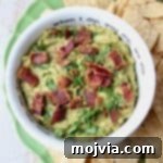 Guacamole in white bowl topped with cooked bacon, with tortilla chips on the side