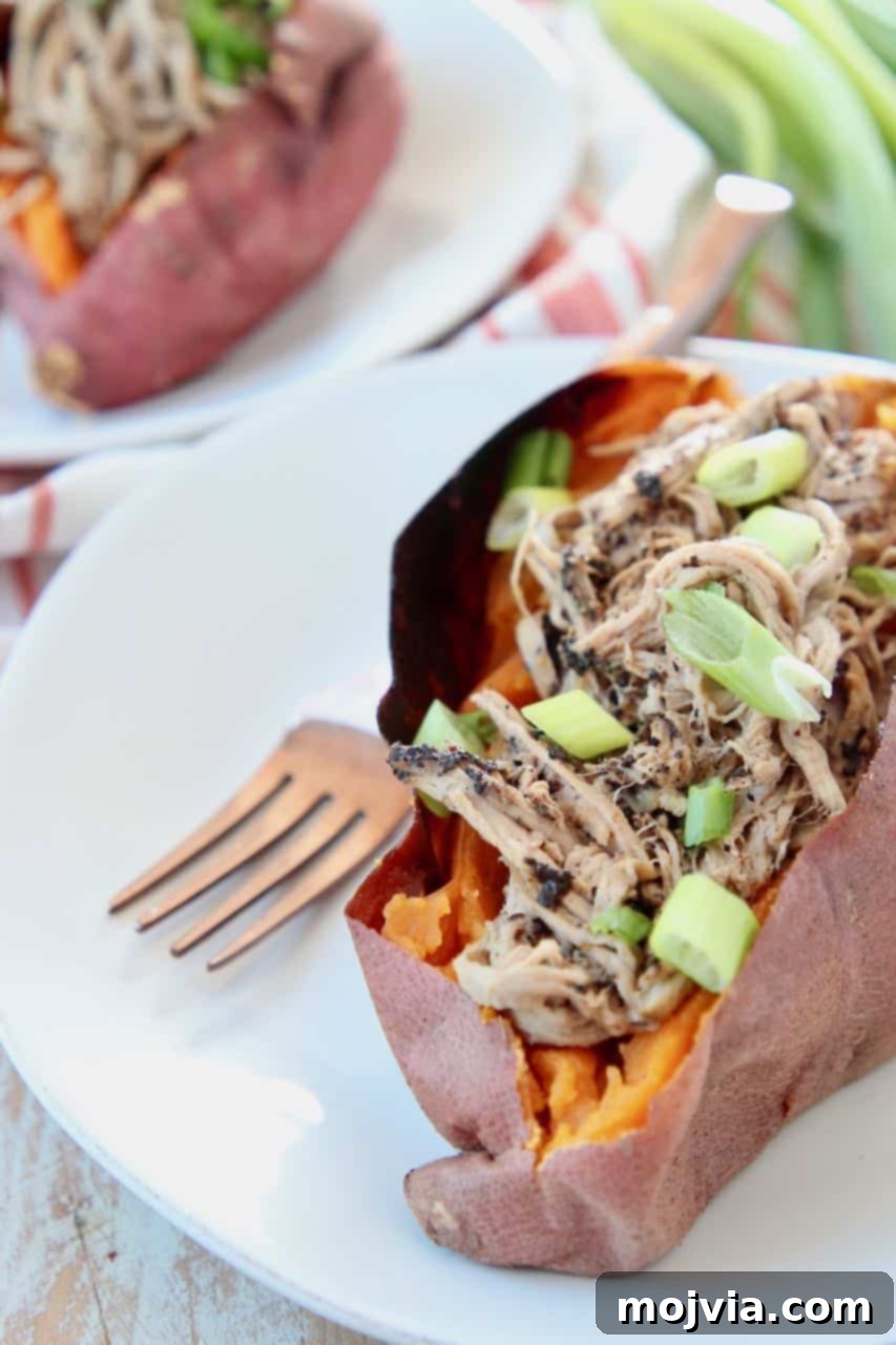 Close-up of fluffy pulled pork tenderloin overflowing from a roasted sweet potato, garnished with vibrant diced green onions, on a white plate with a fork.
