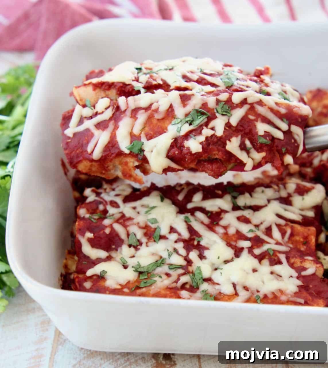 Rustic Mushroom and Squash Enchiladas 3 A single veggie enchilada being lifted from a white casserole dish with a spatula, showcasing the melted cheese and rich red sauce.