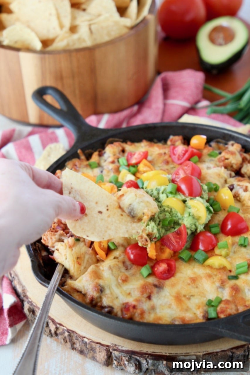 A hand enthusiastically dips a crispy tortilla chip into a serving of hot, cheesy baked taco dip, perfectly garnished with fresh guacamole and diced tomatoes.