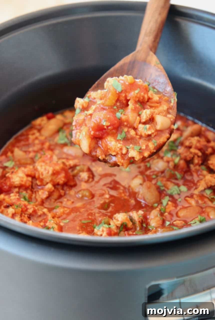 A large serving spoon lifts a generous portion of steaming Buffalo chicken chili from a slow cooker, highlighting its rich texture and chunks of chicken and vegetables.