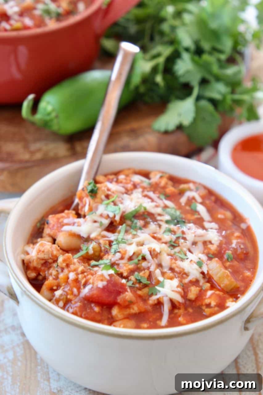 A close-up shot of Buffalo chicken chili in a pristine white bowl, garnished with a single green leaf, accompanied by a spoon, highlighting its inviting texture and rich color.