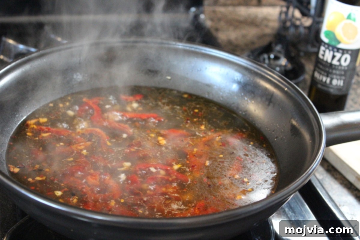 Chicken broth simmering in a skillet with sun-dried tomatoes, garlic, and red pepper, preparing for couscous