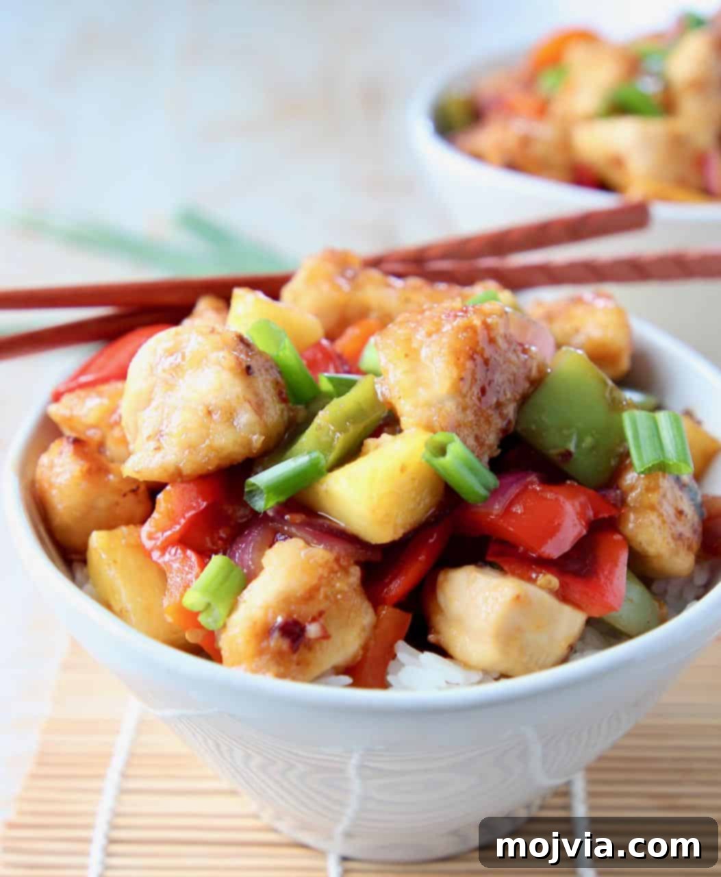 Guilt Free Sweet and Sour Chicken Delight 3 Close-up of golden-brown sweet and sour chicken in a white bowl, garnished with green onions, with chopsticks resting nearby.