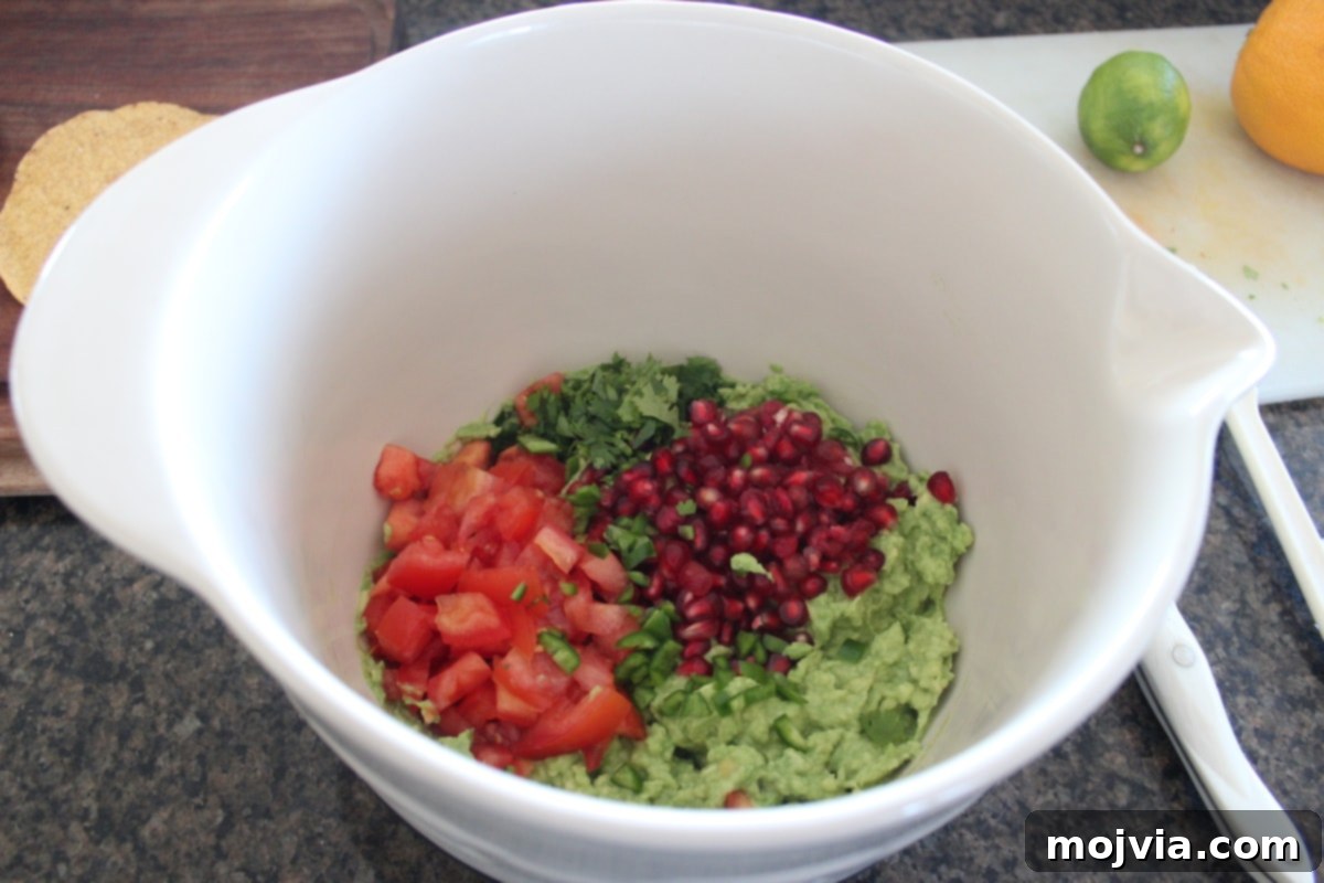 Mashed avocado in a large bowl with diced tomatoes and pomegranate arils being stirred