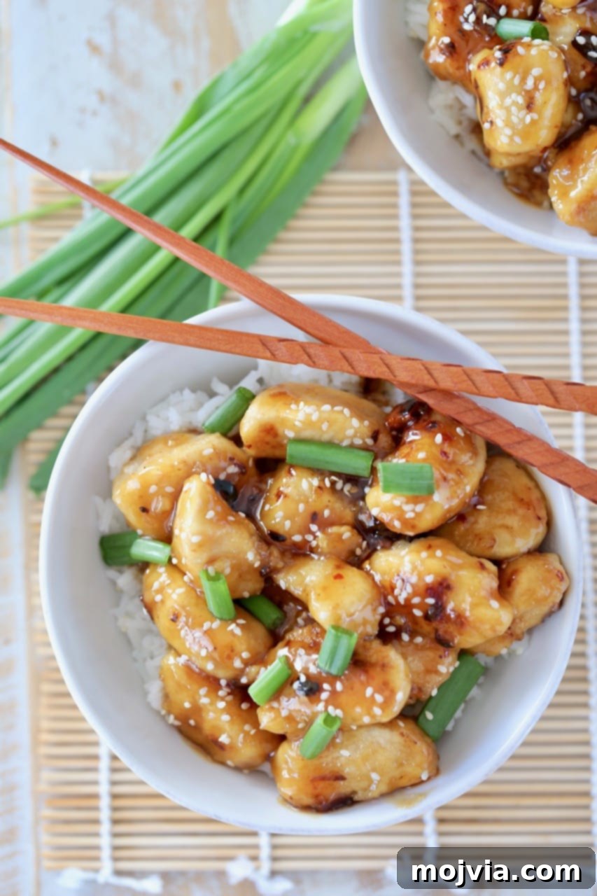 Overhead shot of Chinese honey chicken in white bowl with white rice, green onions and sesame seeds with chopsticks on the side of the bowl and green onions next to the bowl