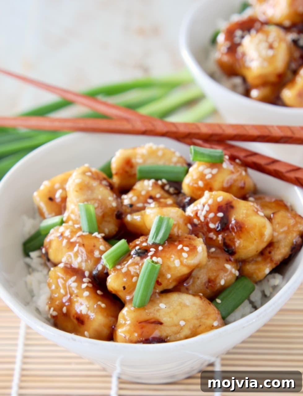 Chinese honey chicken pieces in white bowl with white rice, garnished with green onions and sesame seeds with brown chopsticks on the side of the bowl and whole green onions in the background
