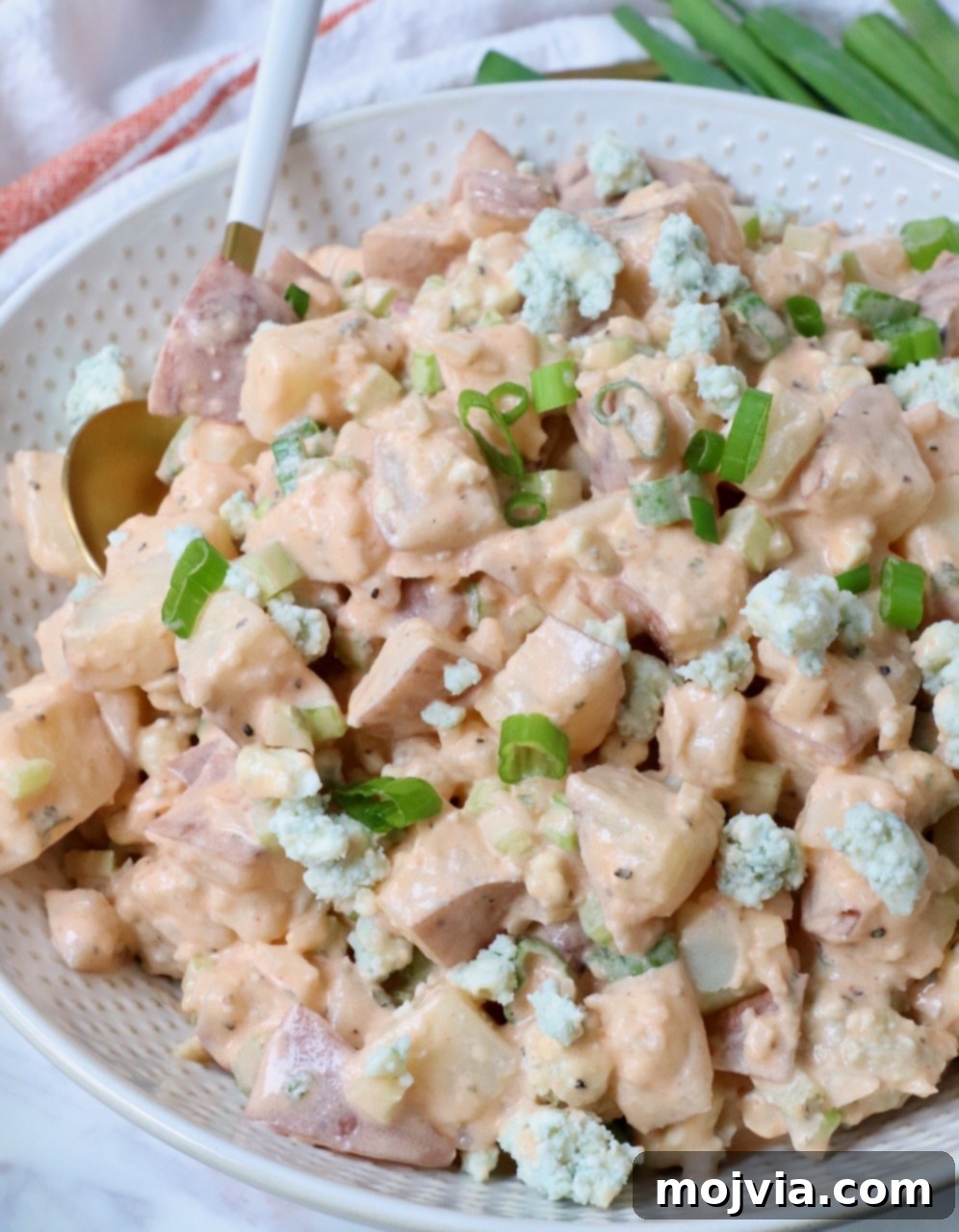 Buffalo Potato Salad in a white bowl, with a serving spoon, ready to be enjoyed. A small side bowl of buffalo sauce adds extra appeal.