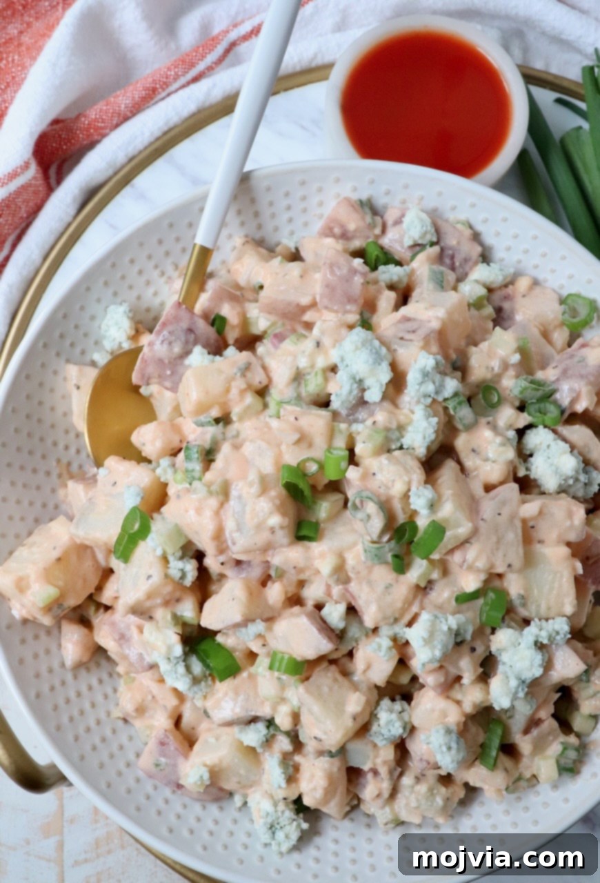 A large bowl of Buffalo Potato Salad with a spoon, alongside a small bowl of buffalo sauce for extra drizzle and garnish.