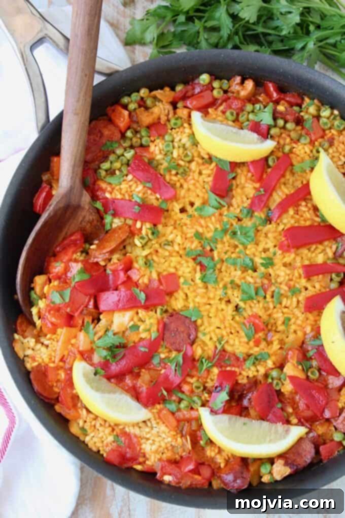 Overhead image of a beautifully cooked chicken and chorizo paella in a skillet with a wooden serving spoon, garnished with lemon wedges.