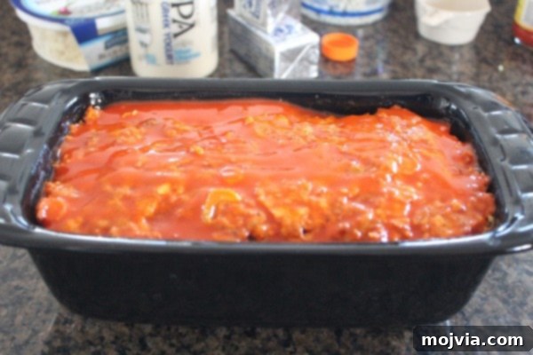 Buffalo sauce brushed on top of a partially baked meatloaf in a loaf pan, adding flavor and moisture.