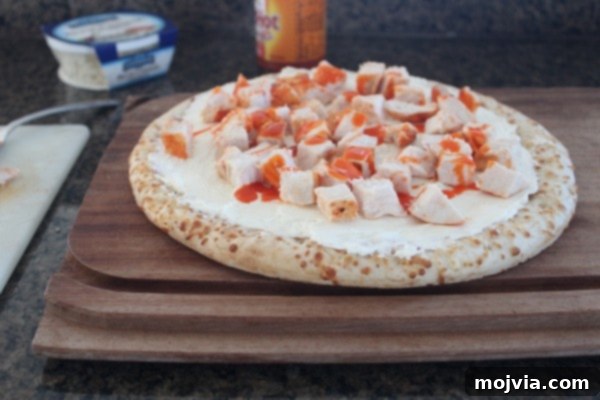 Loaded Buffalo Chicken Pizza 5 A pizza crust topped with a white cheese sauce, diced buffalo chicken, and a generous drizzle of buffalo sauce on a wooden cutting board.