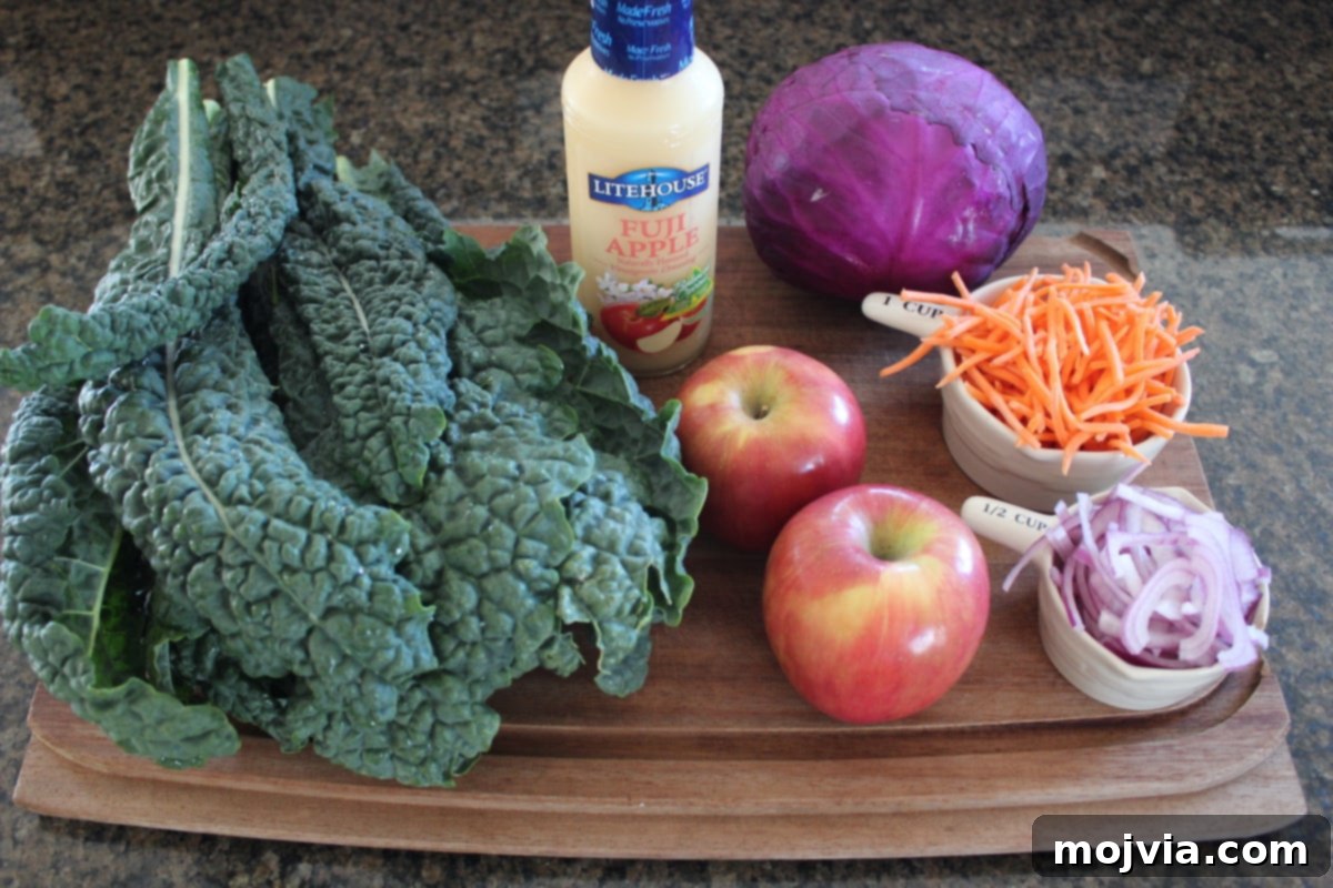 Crisp Kale and Apple Slaw 3 A selection of fresh ingredients for Kale Apple Slaw, including kale, red cabbage, red onion, Fuji apple, and carrots, arranged on a wooden cutting board.