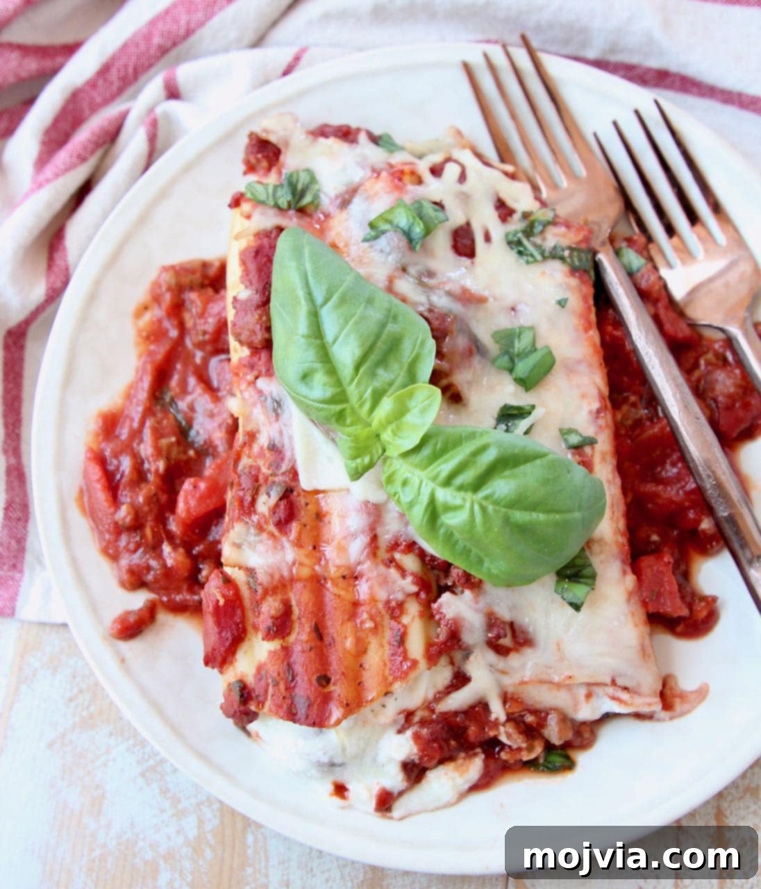 Hearty Three-Cheese Manicotti with Robust Italian Sausage Marinara 5 A serving of manicotti on a plate, garnished with fresh basil leaves, with copper forks beside it.