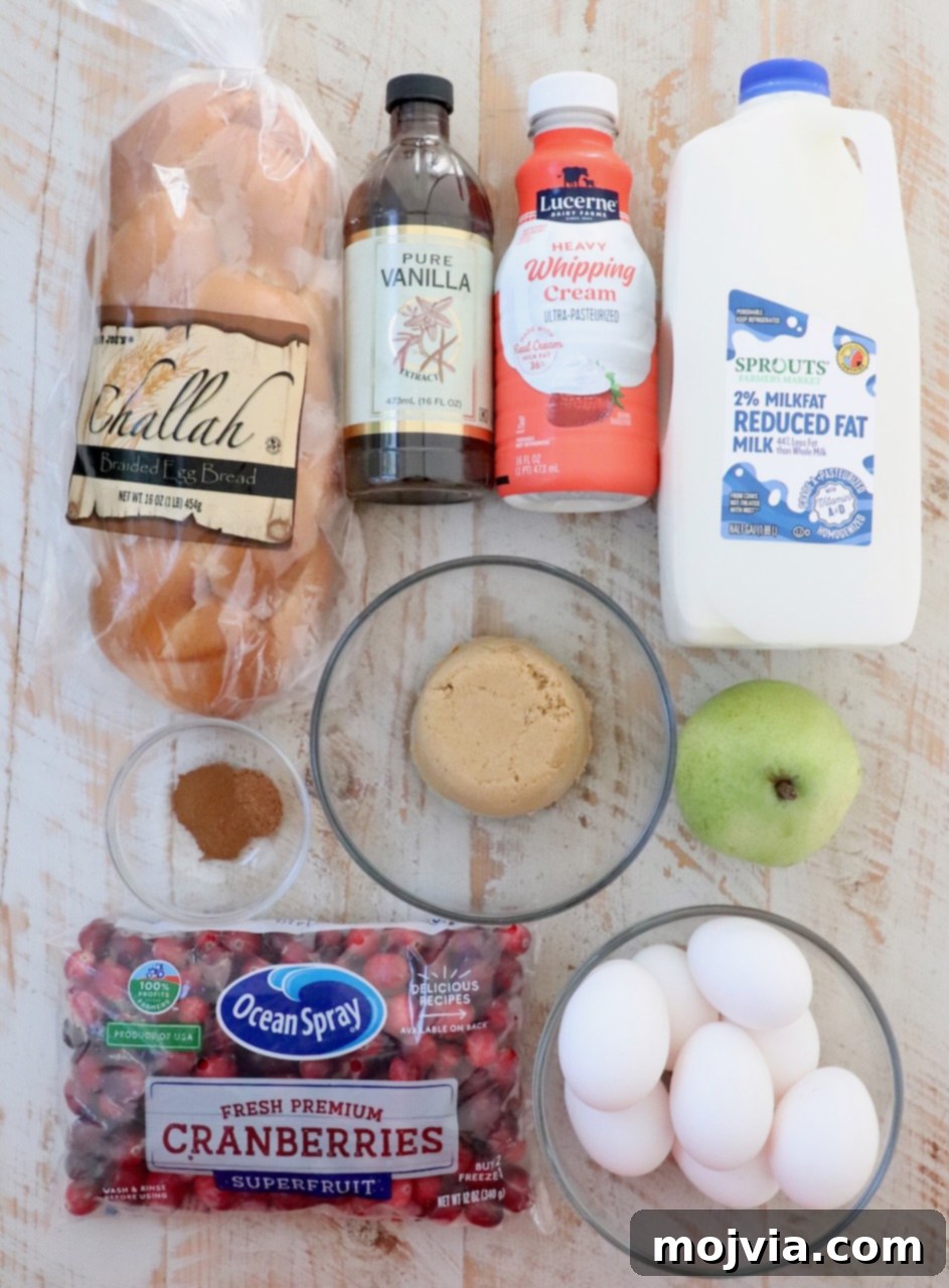 Assortment of fresh ingredients for French Toast Bake, including challah bread, cranberries, pears, milk, eggs, and spices, laid out on a white wooden board.