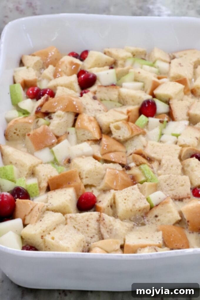 Cubed bread, fresh cranberries, and diced pears soaking in the egg custard mixture in a casserole dish, ready for an overnight refrigeration.