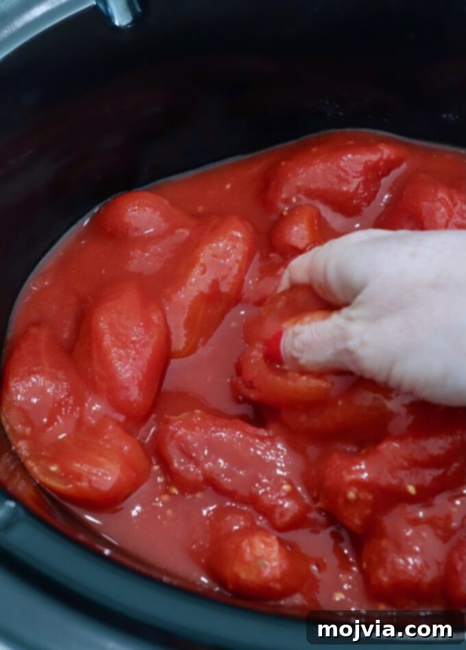 Set-and-Forget Slow Cooker Meatballs and Marinara 4 hand reaching into slow cooker filled with whole canned tomatoes