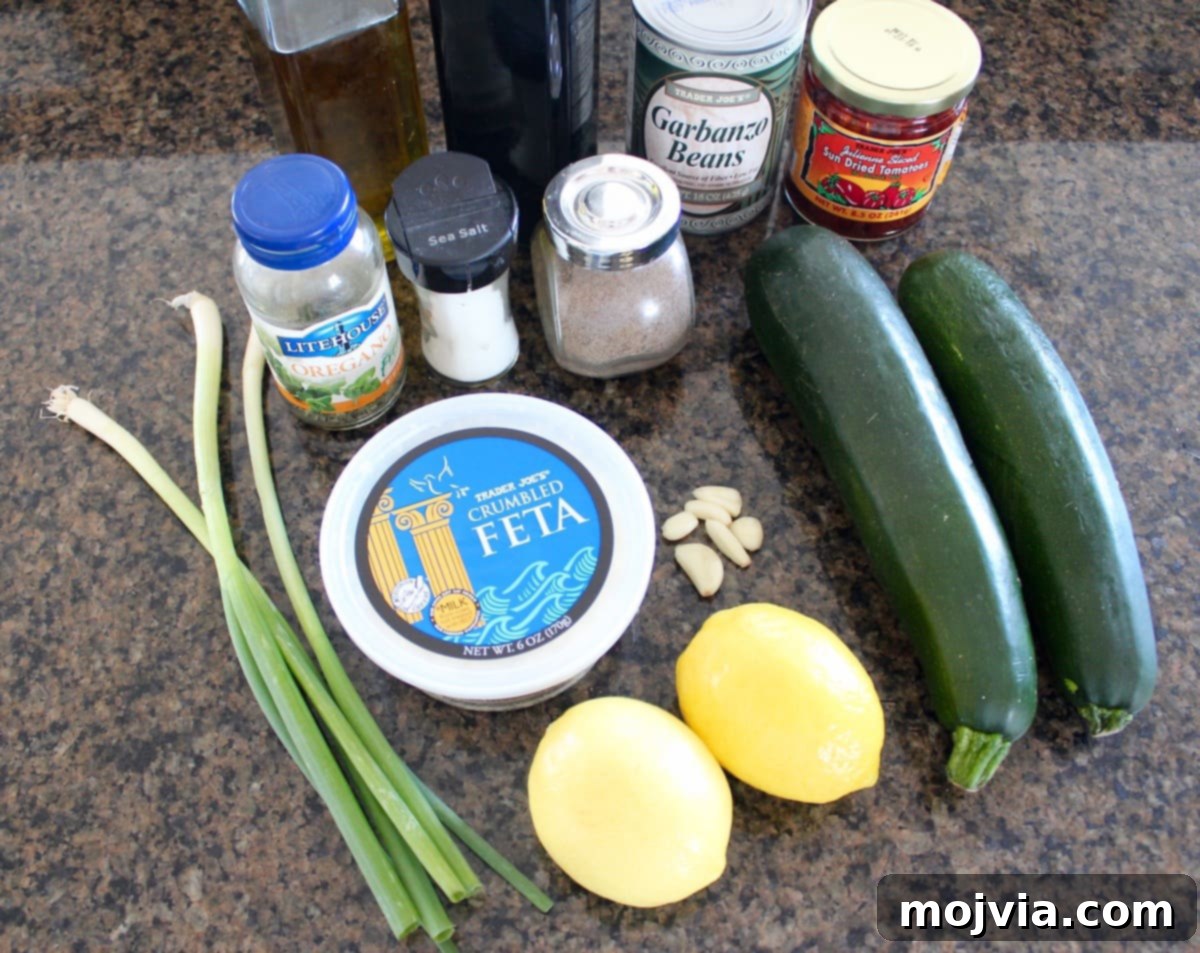 Fresh ingredients for Greek Zucchini Noodles laid out on a granite countertop, including zucchini, chickpeas, sun-dried tomatoes, feta, lemons, and herbs