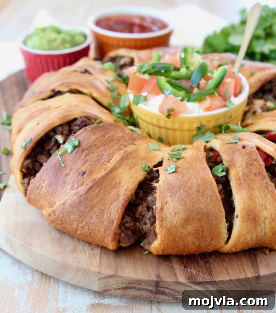Fiesta Taco Ring 2 Golden brown crescent roll taco ring filled with seasoned ground beef and cheese, garnished with fresh cilantro, ready to be served on a wooden cutting board with a side of sour cream and salsa.