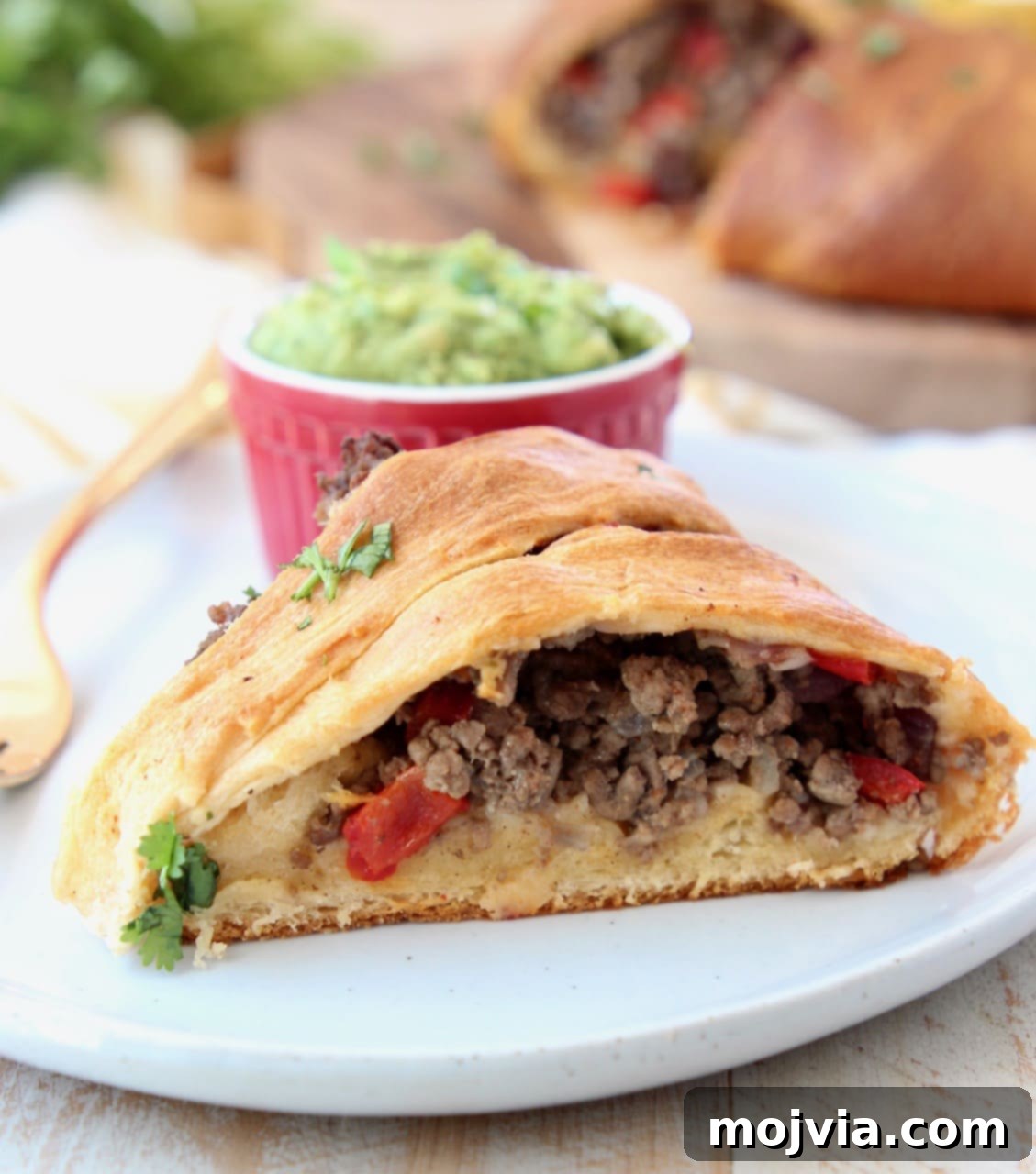 Fiesta Taco Ring 6 A perfectly portioned slice of crescent roll taco ring, showcasing the rich ground beef filling, served alongside a small ramekin of fresh guacamole and a gold fork.