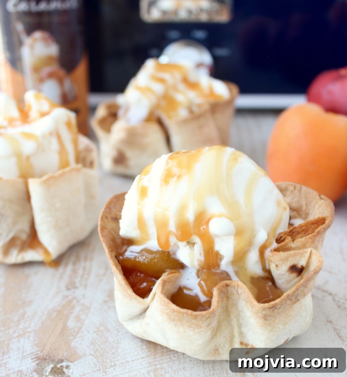 Effortless Fruit-Filled Dessert Shells 2 crispy taco shell cups filled with slow cooked fruit, topped with a scoop of ice cream and caramel syrup on a white wood board in front of a blue crock pot