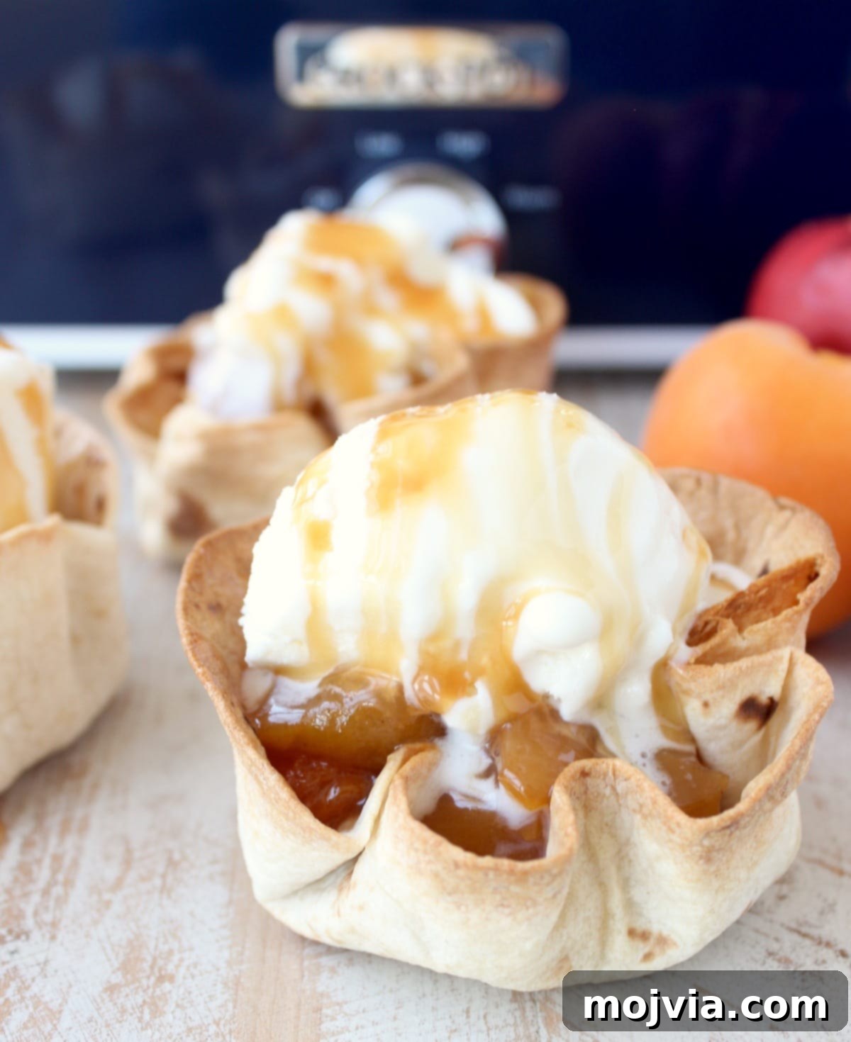 Effortless Fruit-Filled Dessert Shells 9 crispy taco shell cups filled with slow cooked fruit, topped with a scoop of ice cream and caramel syrup on a white wood board in front of a blue crock pot