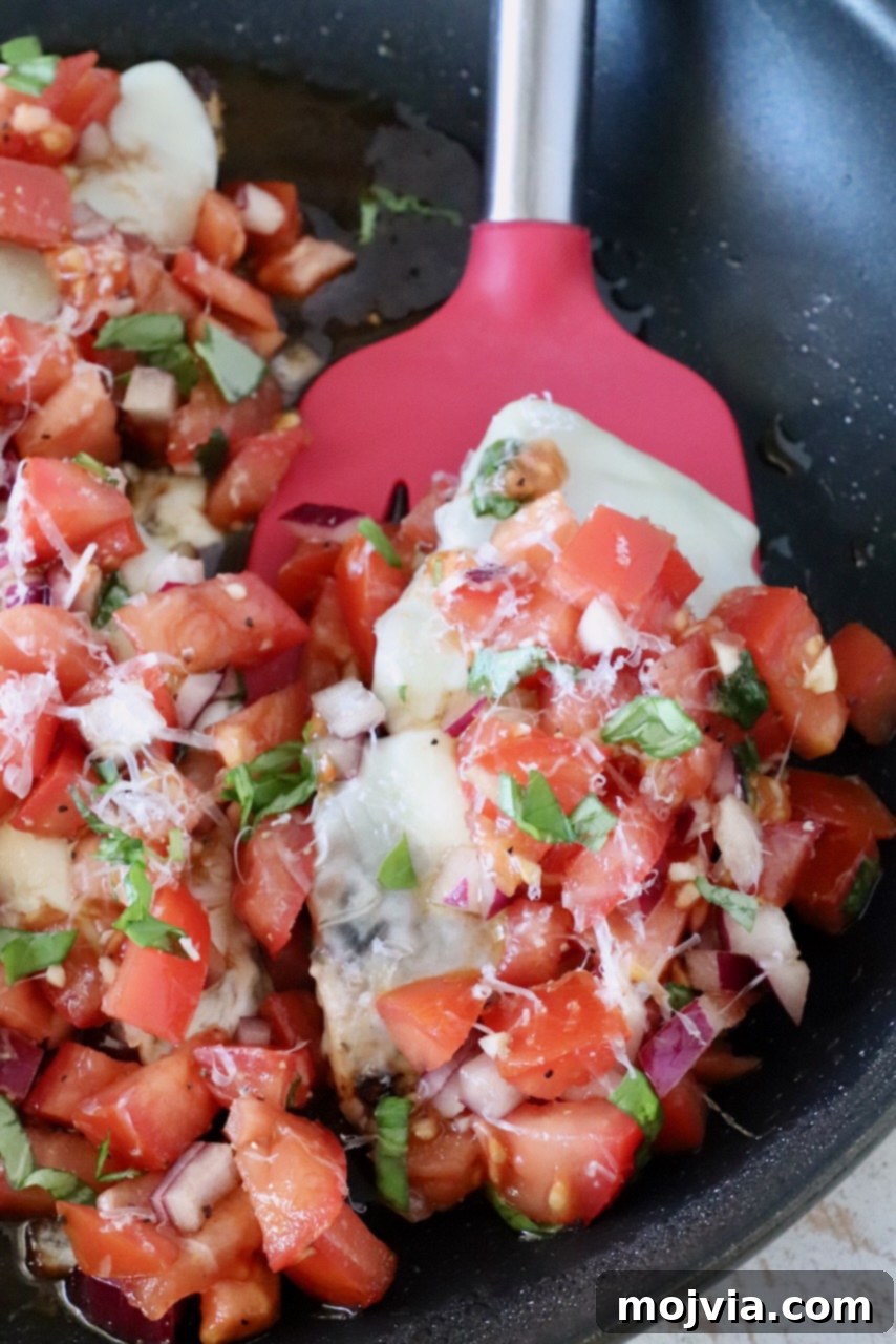 cooked chicken topped with cheese and tomato bruschetta in a skillet with a red spatula