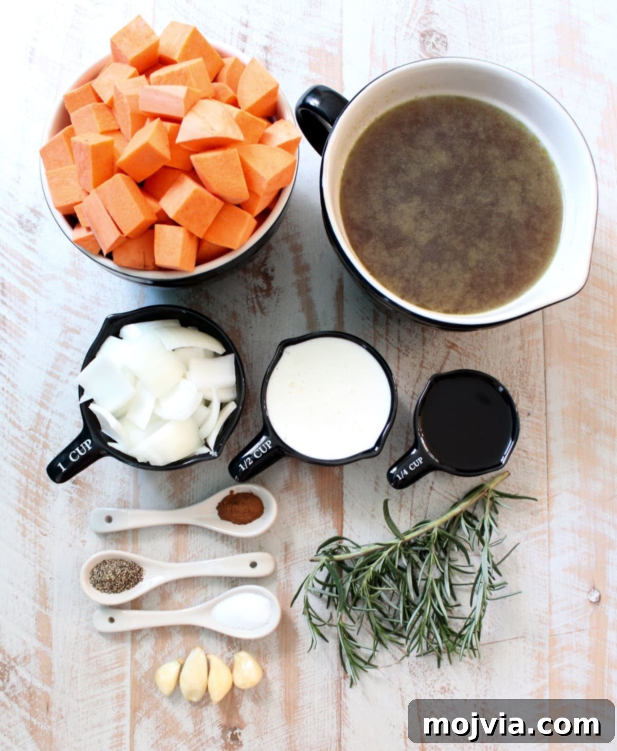 Spiced Sweet Potato Warmth 3 A selection of fresh ingredients for sweet potato soup, including whole sweet potatoes, an onion, garlic cloves, and fresh rosemary, arranged on a white wooden board.