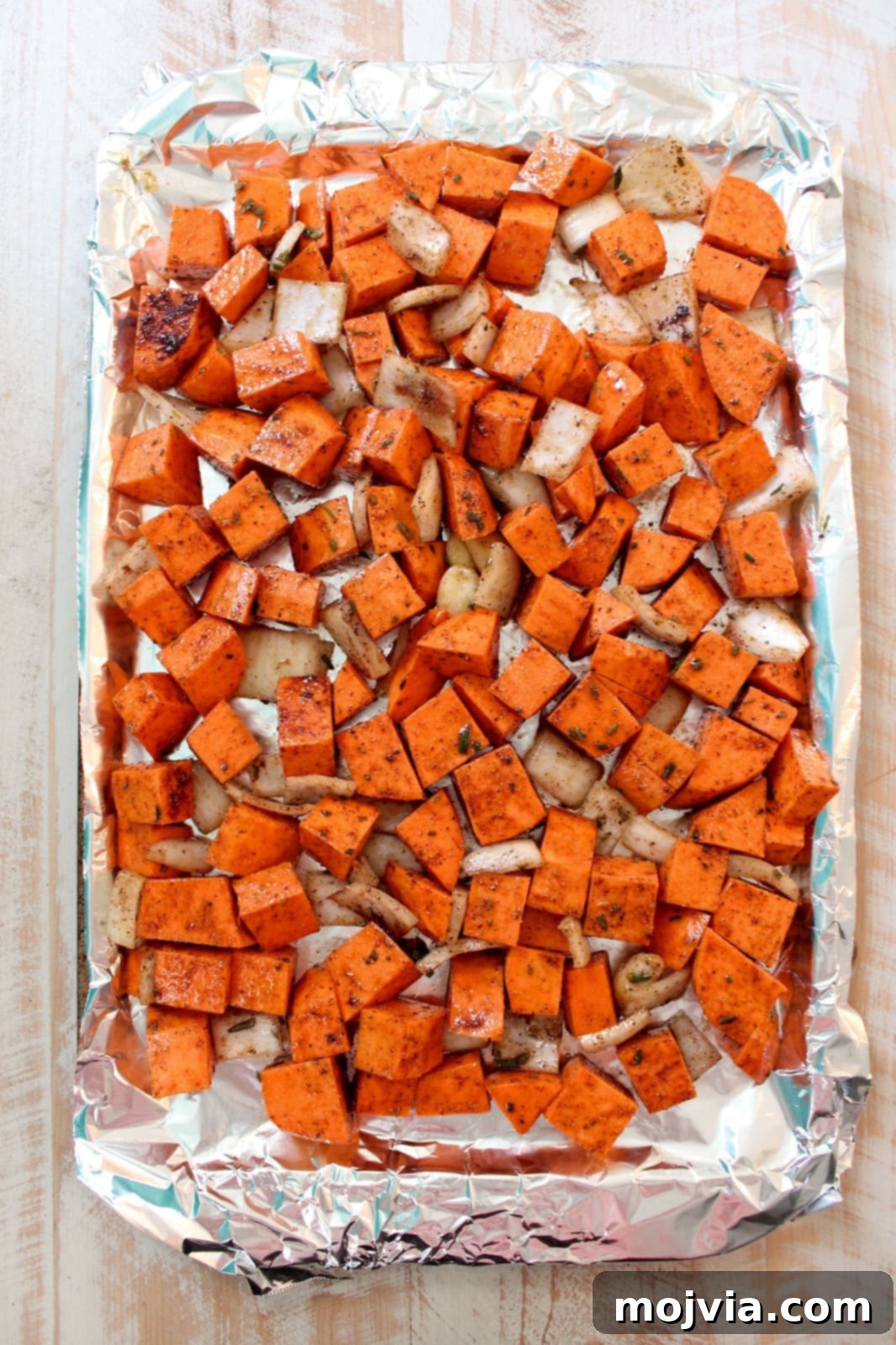 Spiced Sweet Potato Warmth 4 Cubed sweet potatoes, diced onion, and whole garlic cloves seasoned with herbs and olive oil, spread evenly on a foil-lined baking sheet, ready for roasting.