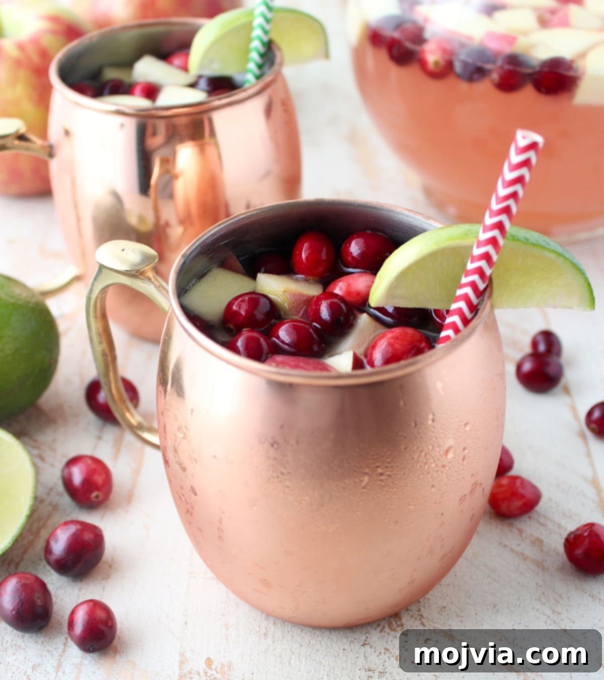 Cranapple Moscow Mule Punch 2 Copper mugs filled with Cranberry Apple Moscow Mule Punch, garnished with fresh cranberries and diced apples, set against a festive background.